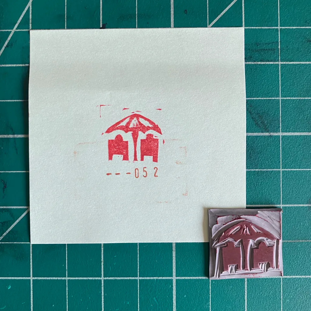 A white sheet displays a red outline drawing of a pitched-roof structure and the number -- -052, next to a small, reddish-brown, textured, three-part replica of the design.