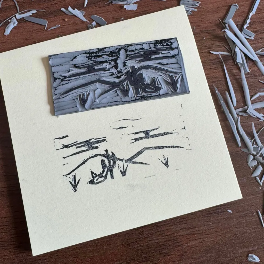 A pale yellow paper holds a rectangular, dark, highly textured element above a grouping of abstract, black, angular lines.