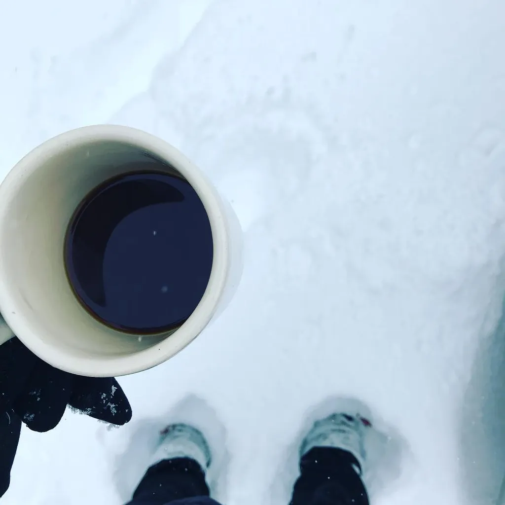 Overhead view showing two white mugs filled with dark liquid and a pair of boots positioned against a vast, bright white expanse of snow.