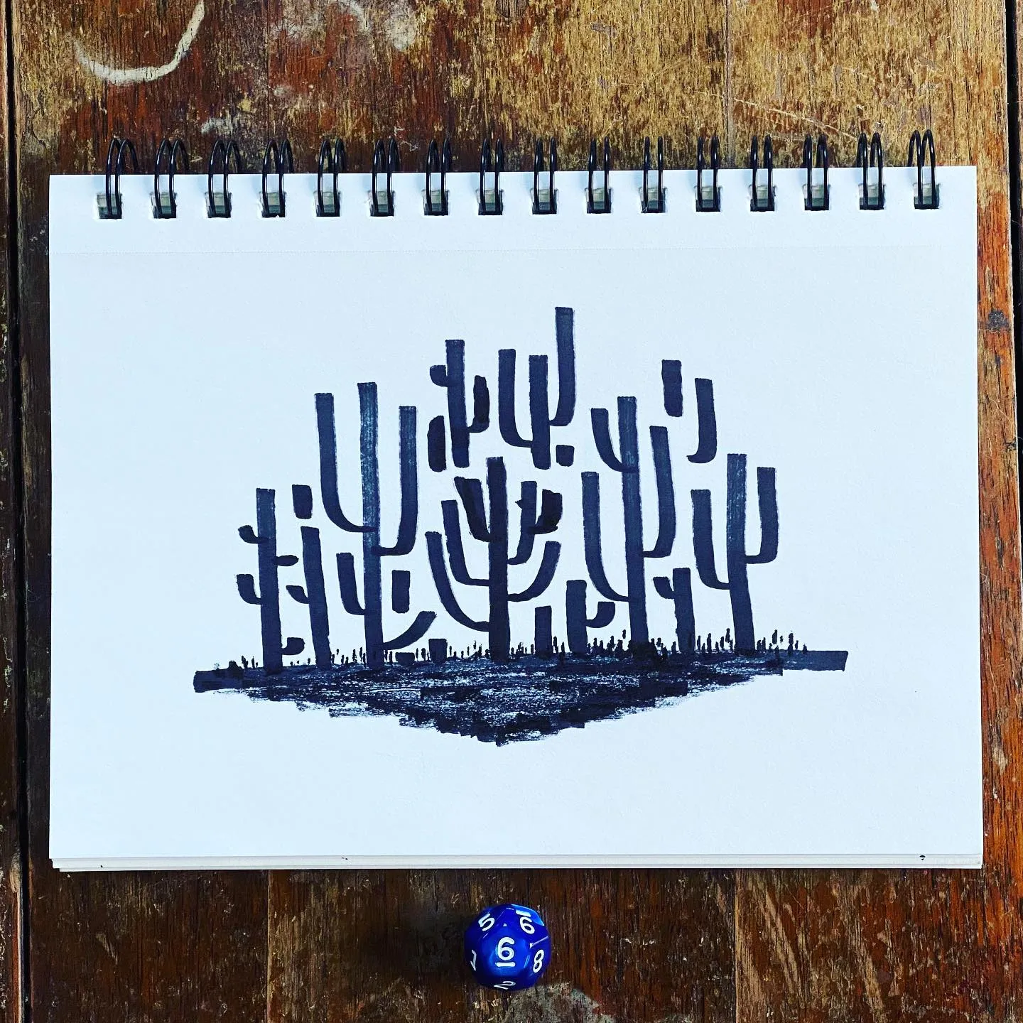 A black drawing of stylized, branching desert plants sits on white paper, which rests on a dark, textured wooden surface next to a blue and white dice.