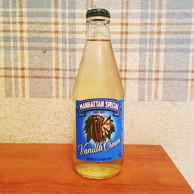 A glass bottle containing pale yellow liquid rests on a reddish-brown wooden surface, set against a plaid background of beige and brown stripes, with a label featuring dark text and a stylized animal head.