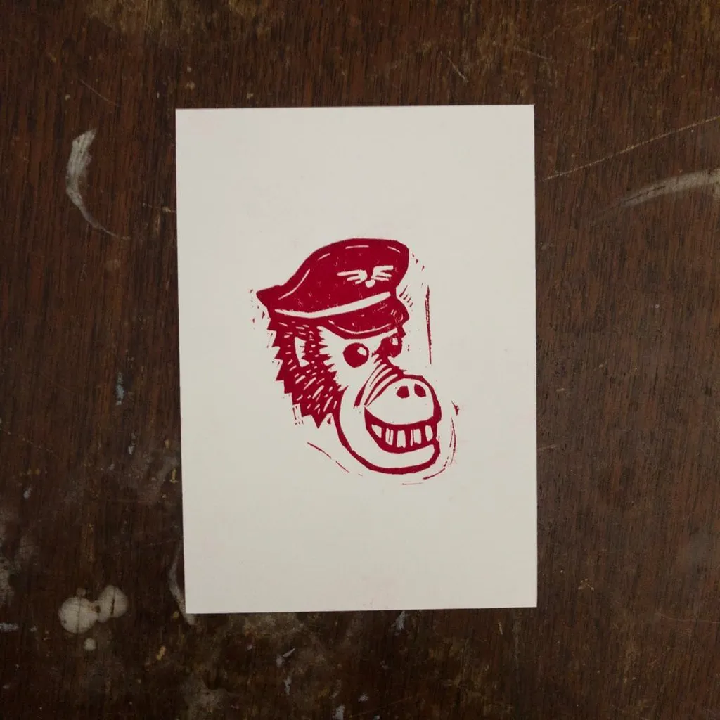 A stylized, red drawing of a monkey head wearing a peaked cap, centered on a small white card placed on a dark, textured wooden background.