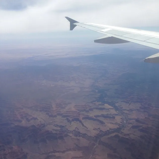 A gray airplane wing extends across the frame, overlooking a vast, arid landscape of browns and tans that meets a hazy white sky and low clouds.