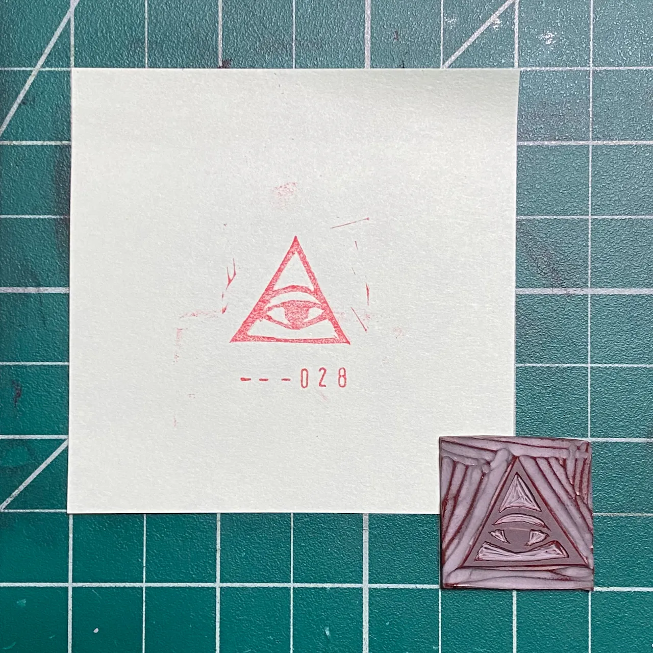 A white paper on a green grid background displays a red drawing of an eye within a triangle and the number --028, next to a small, reddish-brown square featuring a geometric triangular pattern.