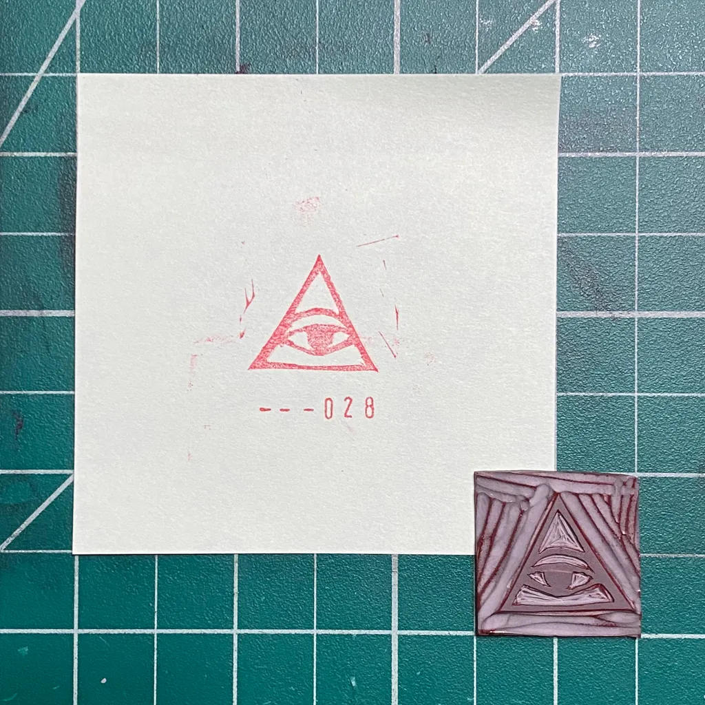 A white paper on a green grid background displays a red drawing of an eye within a triangle and the number --028, next to a small, reddish-brown square featuring a geometric triangular pattern.