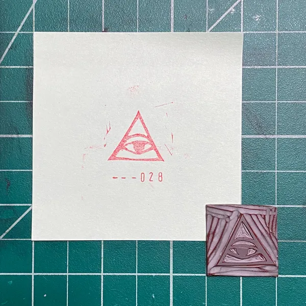 A white paper on a green grid background displays a red drawing of an eye within a triangle and the number --028, next to a small, reddish-brown square featuring a geometric triangular pattern.