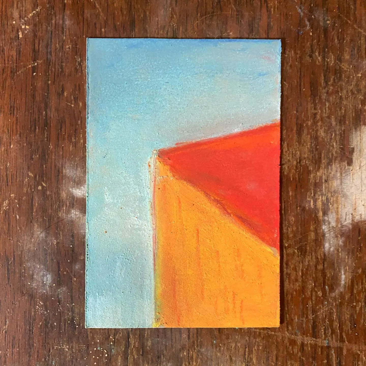 A rectangular painting on a wooden surface shows a light blue field meeting a large orange and red diagonal shape, which is set against a dark brown wood grain background.