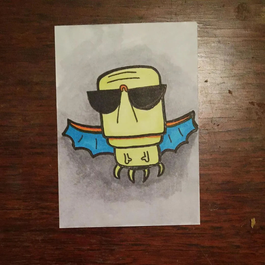 A stylized yellow creature with large black sunglasses and outstretched blue wings is drawn on white paper, surrounded by pencil shading, placed on a dark wooden surface.