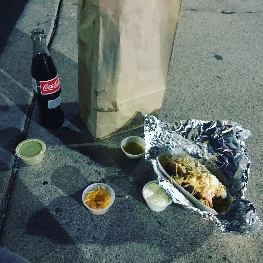 A collection of takeout items rests on rough concrete, including a dark soda bottle, a large brown paper bag, several small dipping cups, and a piece of fried food in crumpled silver foil.