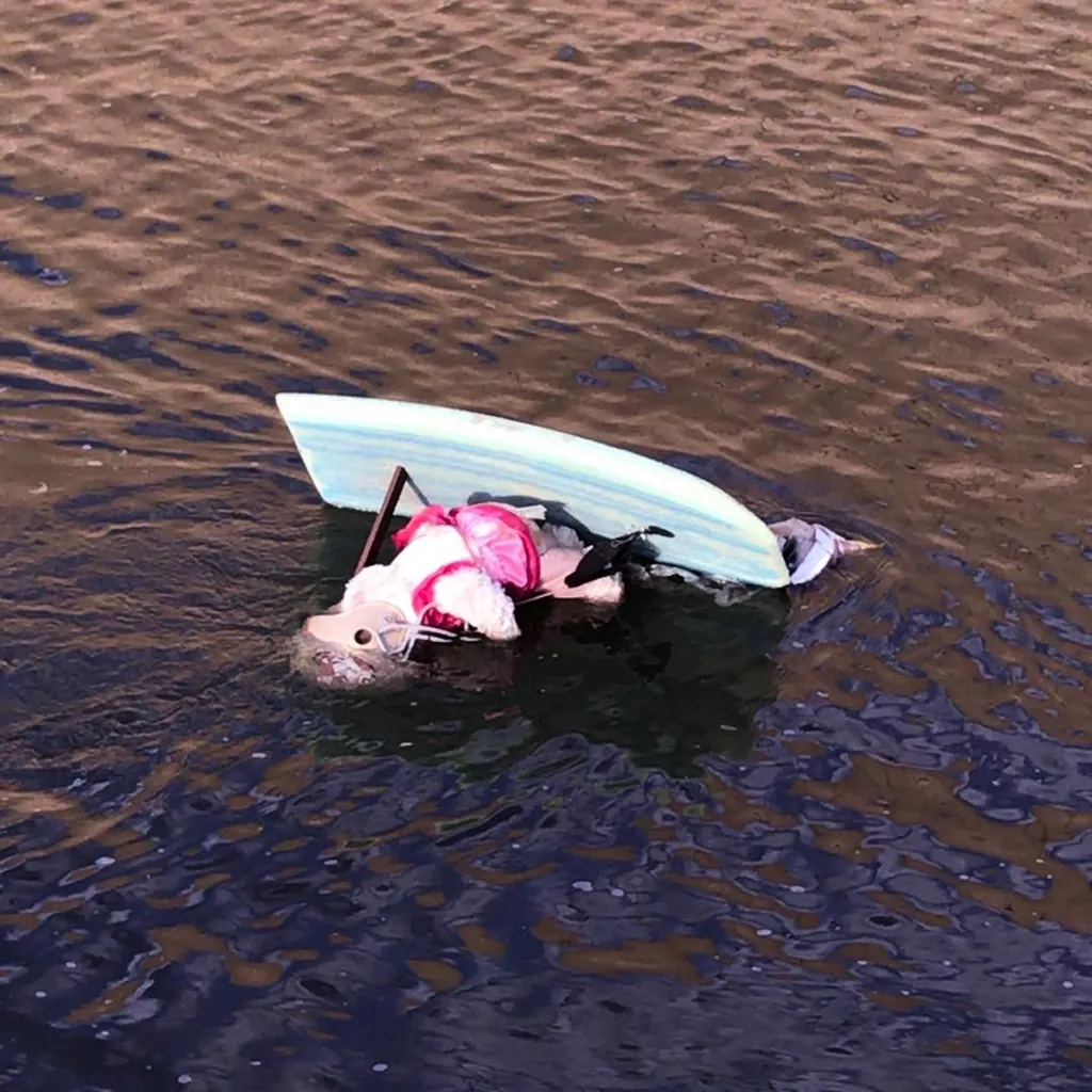 Teal and dark surfboards float in rippling water, carrying a pink and black object, with the water surface transitioning from light brown to deep black.