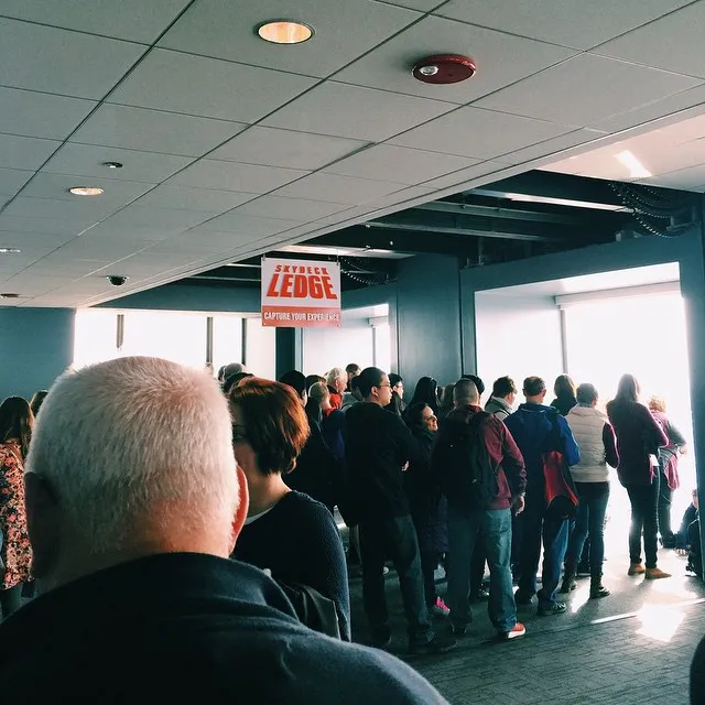 The composition shows the back of a man's head in the foreground, looking over a crowd of people gathered in a large, modern indoor space toward a bright exit.