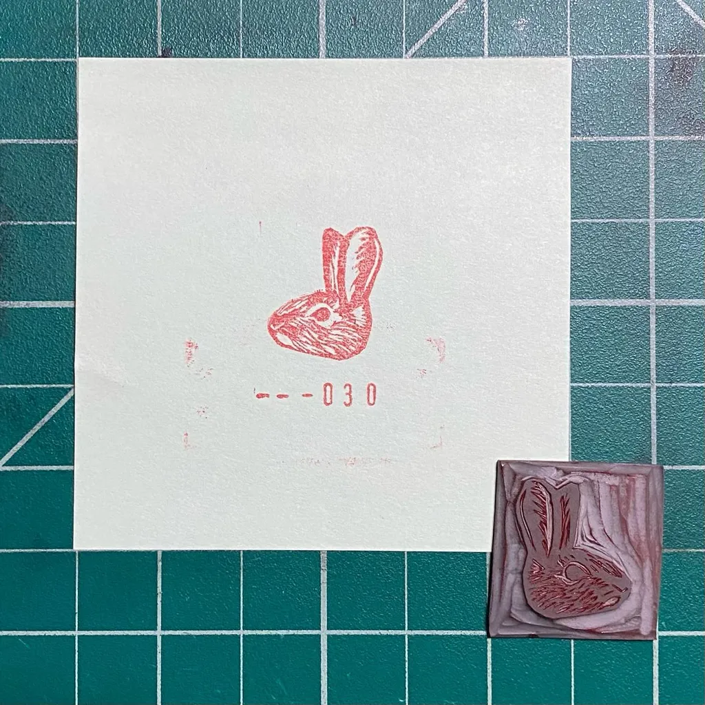 A reddish sketch of a rabbit and the number 030 are on white paper, placed on a teal grid next to a small, square, reddish-brown print of a rabbit.