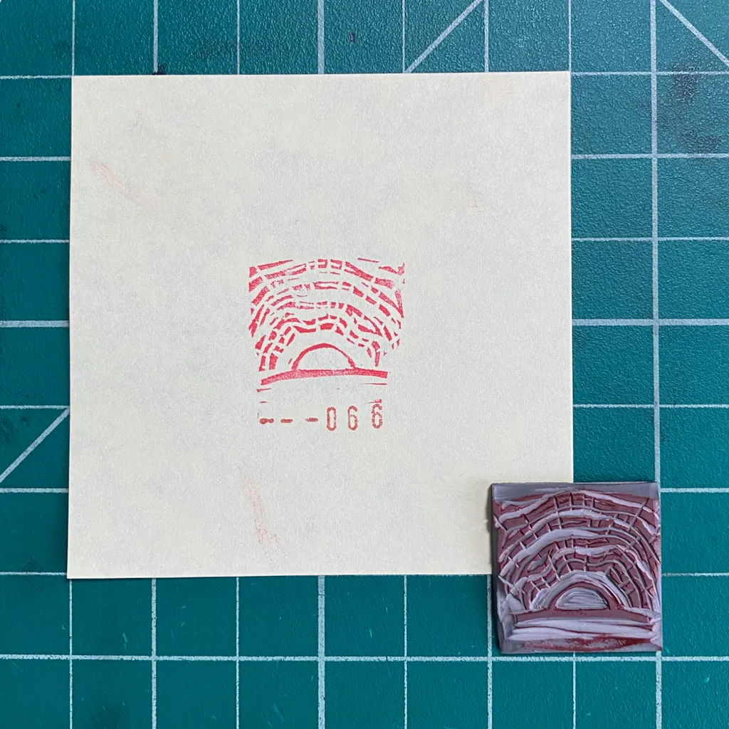 A white square paper on a teal grid features a red graphic of concentric semi-circles and the numbers --066, next to a small, reddish-brown square with a raised, textured version of the same design.