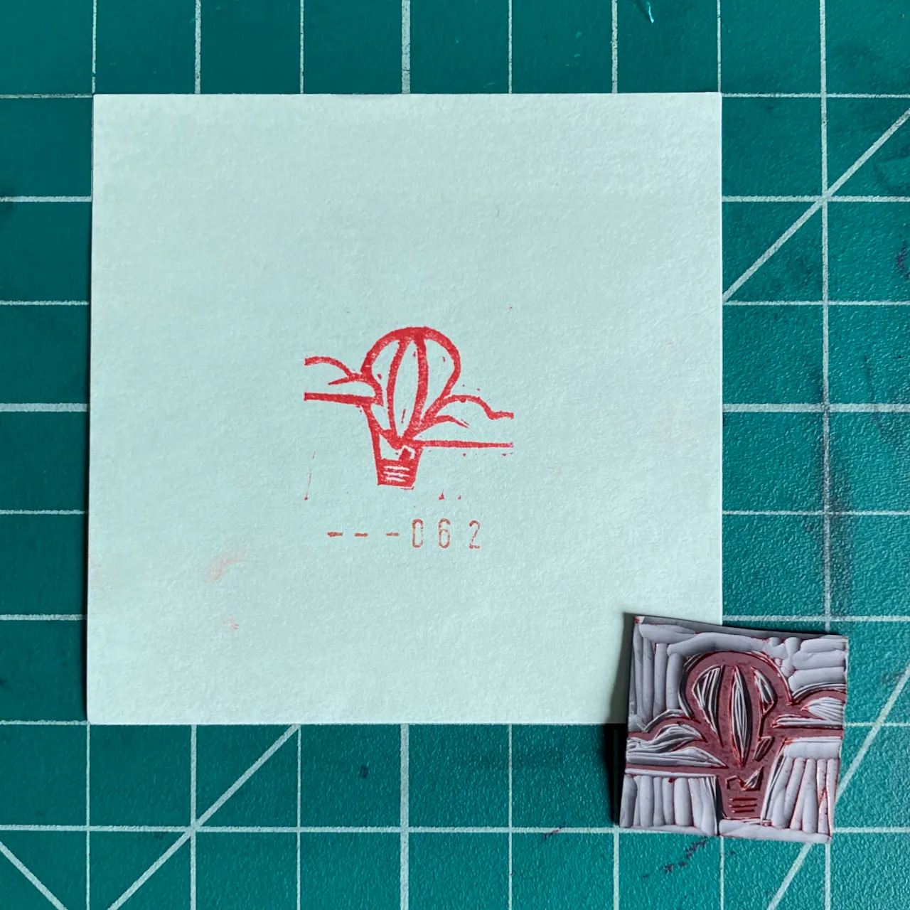 On a green grid background, a light square displays a red drawing of a stylized plant and the number --062, next to a smaller, textured square artwork in shades of red and pink.