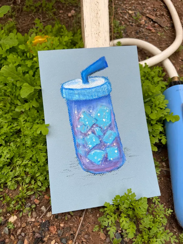 A drawing on light blue paper shows a jar filled with icy blue liquid and chunks of ice, topped with a blue straw, rendered with visible colored pencil marks.