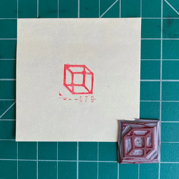 The composition features a cream paper with a red outline drawing of a cube and the number -079-, next to a small, textured square object in pink and white, all set on a green grid.