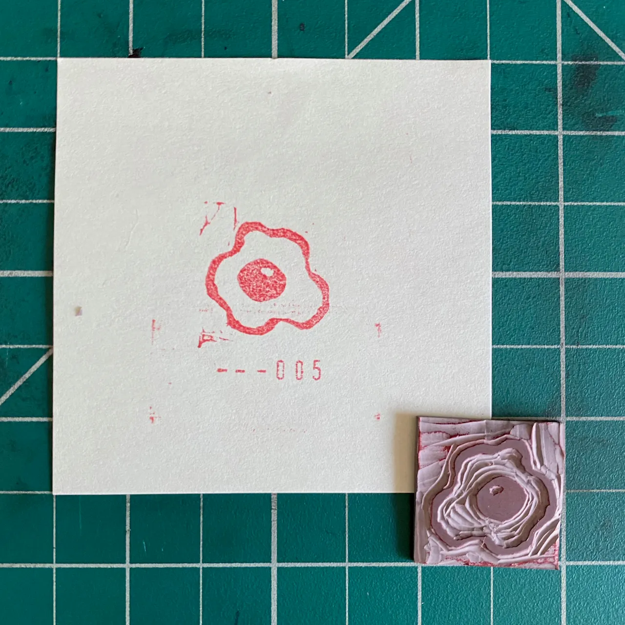 On a teal grid background, a cream paper features a red outline of a stylized flower and the number --005, next to a small, square, pink object with a detailed, layered floral texture.
