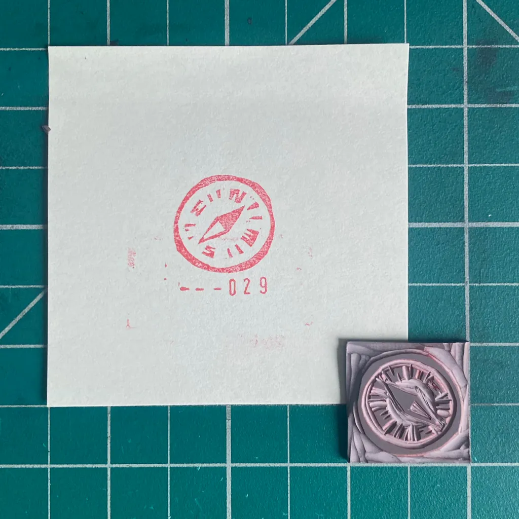 A white square paper with a red circular stamp and the number -029 rests on a teal grid background, next to a small, square, pink and purple embossed object.