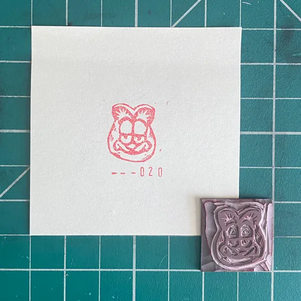 On a teal grid background, a white card holds a pink line drawing of a cat face and the numbers -020, next to a small, mauve, embossed square with a matching cat face design.