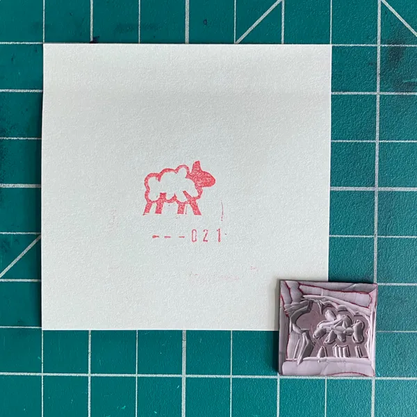 On a teal grid background, a white paper features a red drawing of a sheep and the numbers --021, next to a small reddish square print showing a group of figures.