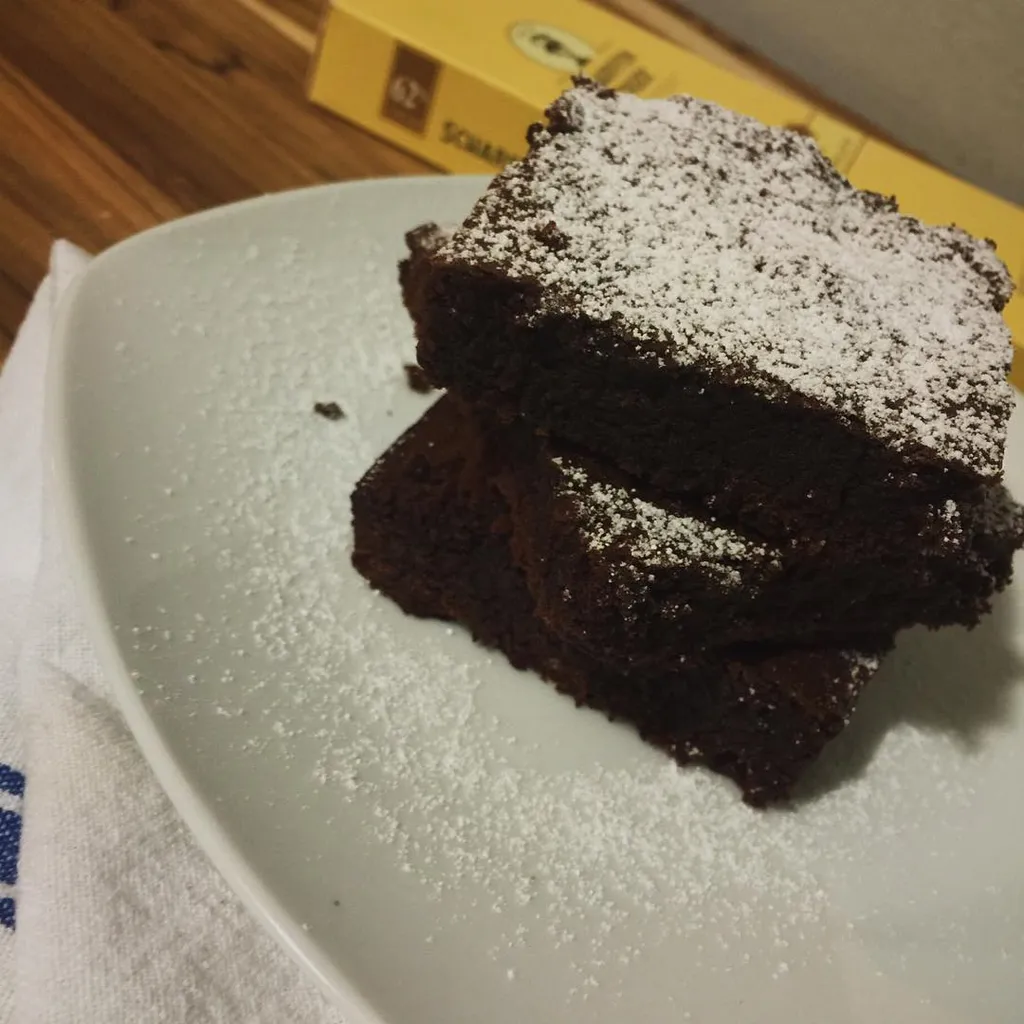 A stack of dark brown, square baked goods, heavily dusted with white powdered sugar, rests on a light ceramic plate in front of a yellow and brown cardboard box.