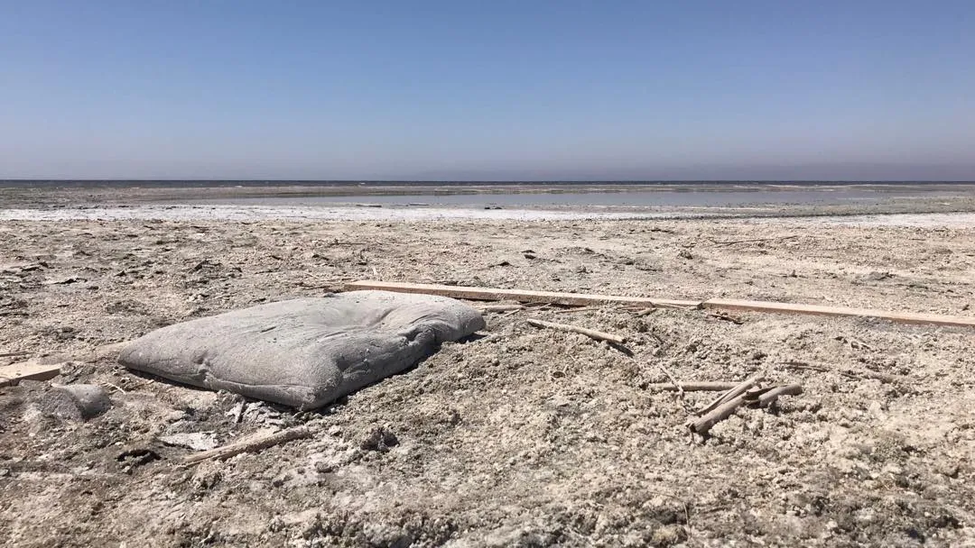 A vast, pale landscape under a clear blue sky features rough, dusty earth, scattered sticks, and a smooth, light rock in the foreground, crossed by a straight debris line.