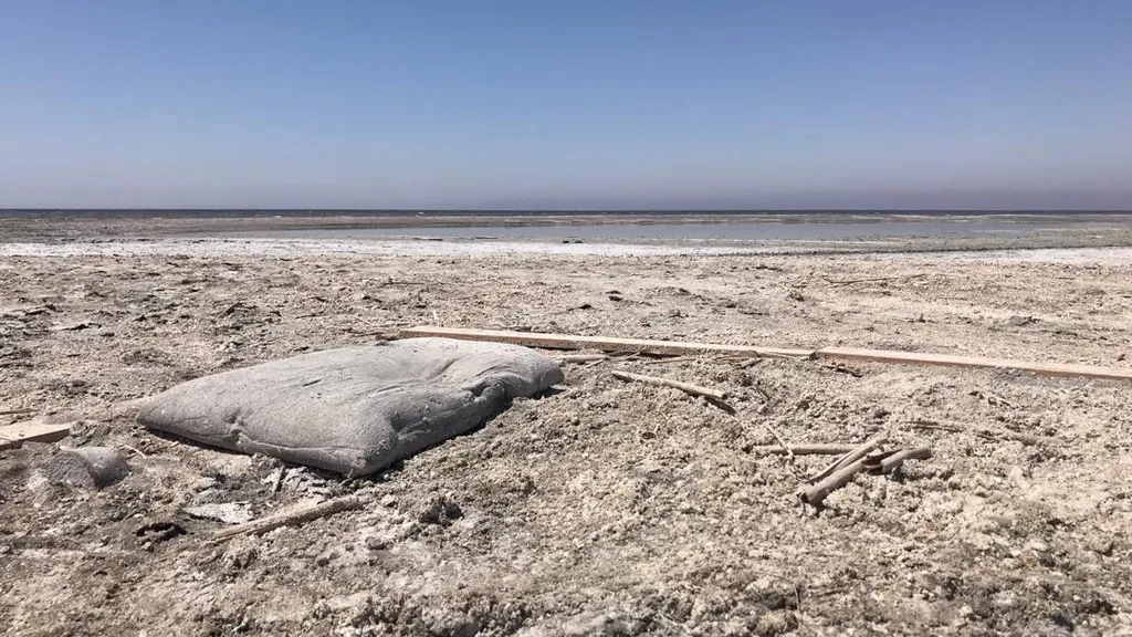 A vast, pale landscape under a clear blue sky features rough, dusty earth, scattered sticks, and a smooth, light rock in the foreground, crossed by a straight debris line.