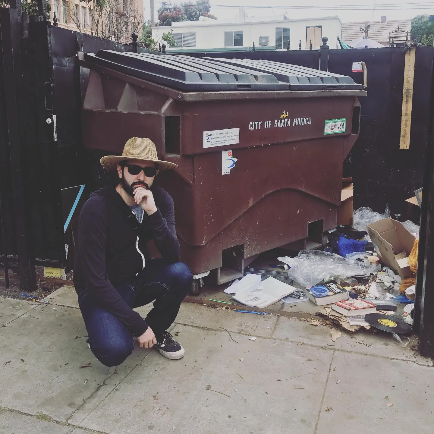 A man in a tan hat and dark clothing kneels on a sidewalk beside a large, brown dumpster marked with white lettering, surrounded by scattered trash.