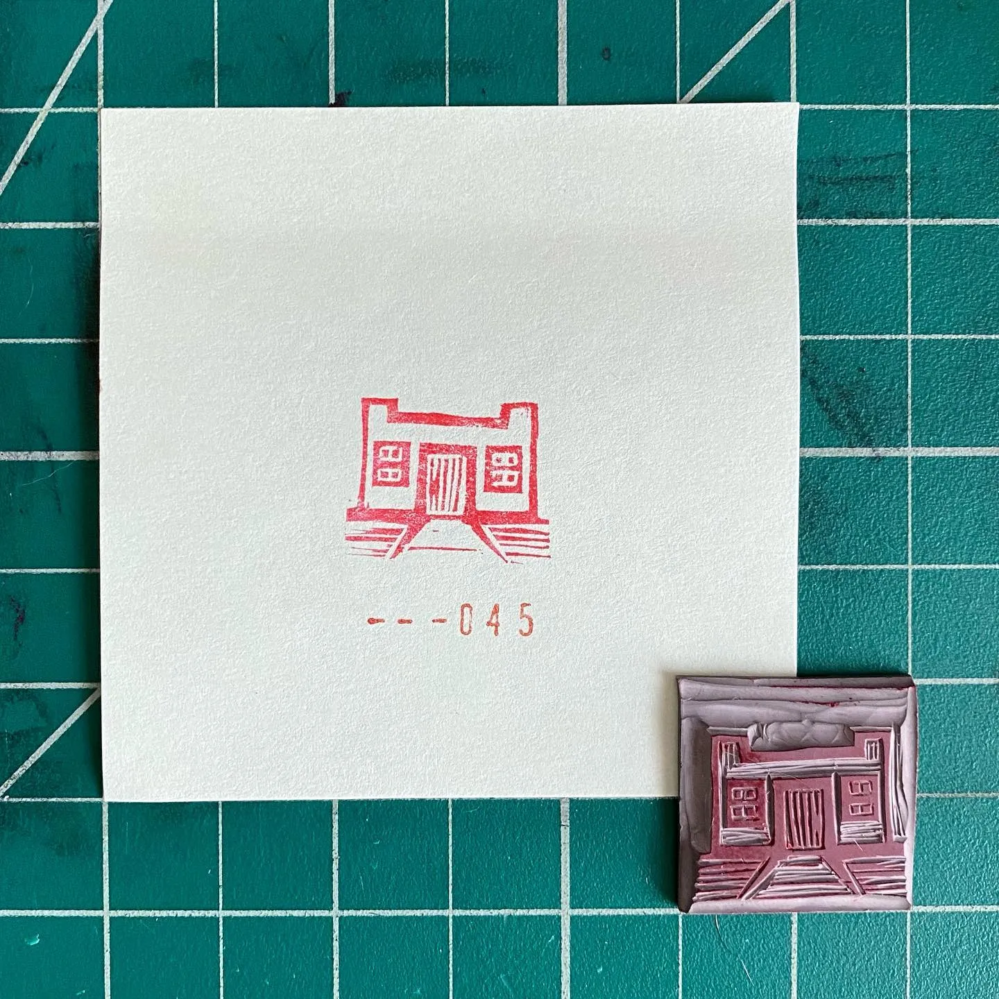 A white card on a teal grid features a red, symmetrical drawing of a building and the number 045, accompanied by a small, textured red square showing a similar structure.