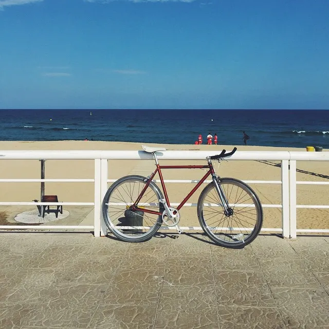 A red bicycle rests on gray pavement, framed by a white railing, with a wide view of the blue ocean and sky beyond the sandy beach.