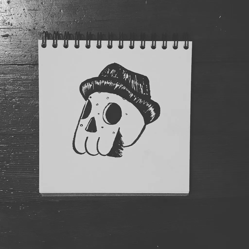 A black and white drawing of a stylized skull wearing a striped hat, centered on a small spiral notepad resting on a dark, textured wooden surface.