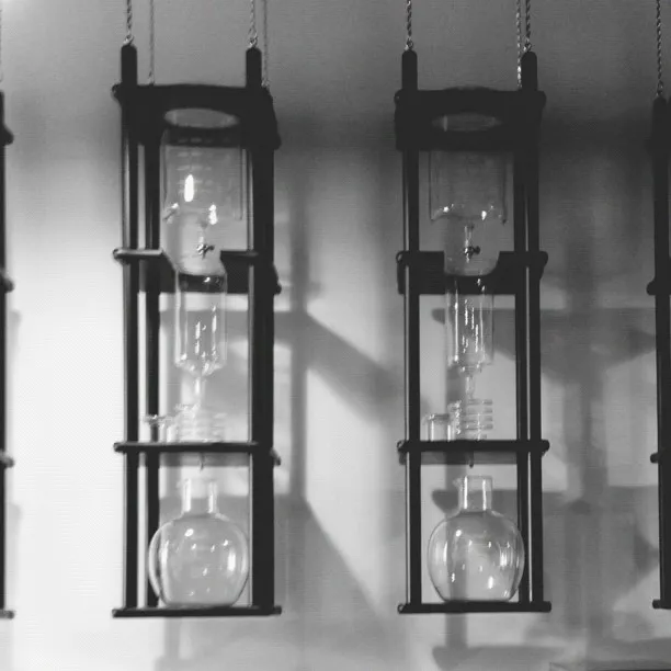 Two hanging, dark rectangular frames suspend stacked glass vessels and apparatuses, set against a plain light background with visible shadows.