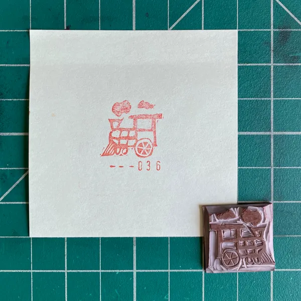 On a green grid background, a white paper displays a red outline drawing of a steam train and the number --036, next to a small, brown, carved object depicting a detailed building facade.