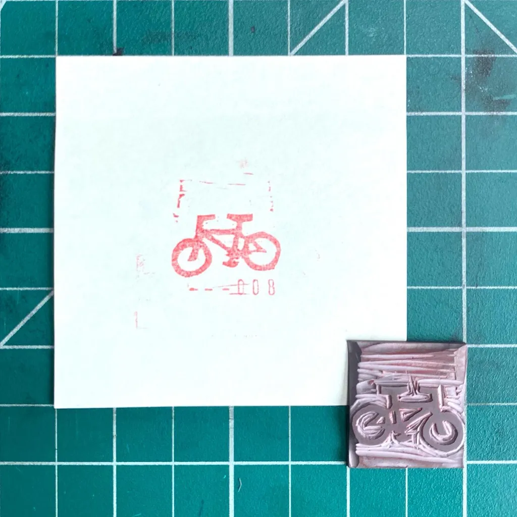 On a teal grid, a white paper displays a simple red bicycle drawing and the number 08, next to a small, textured piece featuring a raised red bicycle design.