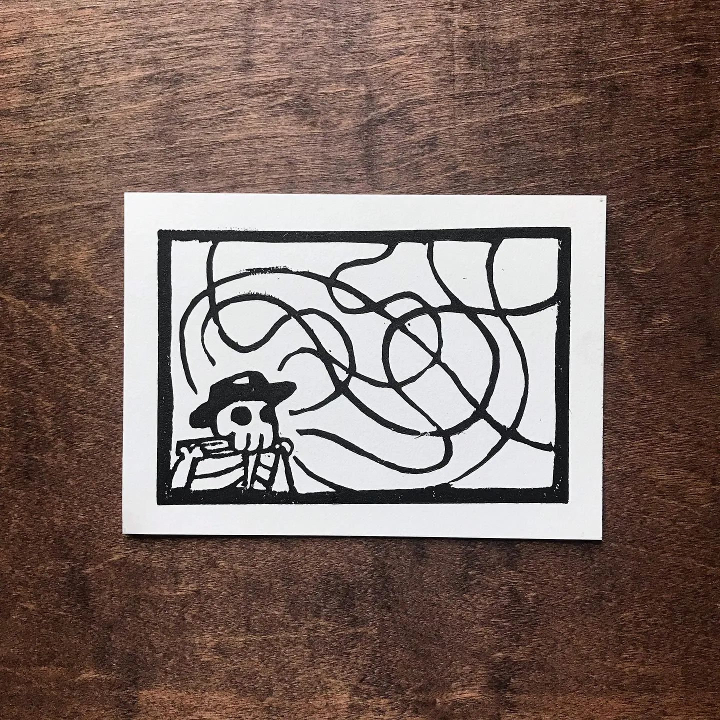 A black and white drawing within a square border features a stylized skull wearing a hat on the left, contrasted by thick, abstract, looping black lines that fill the right side.