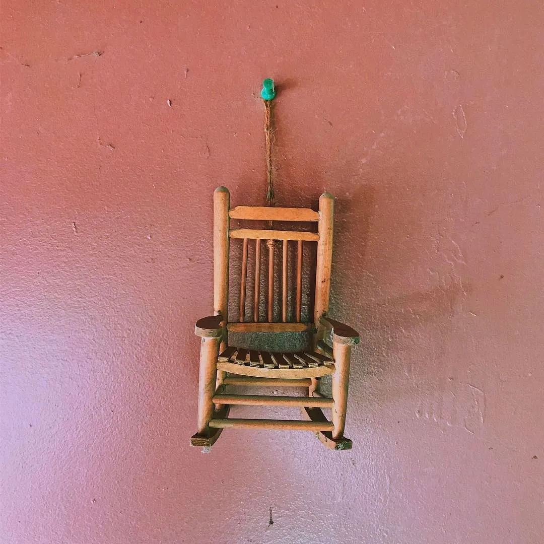 A rustic wooden chair, featuring vertical slats and a slatted seat, hangs centered against a textured dusty rose wall, suspended by a thin cord from a small green hook.