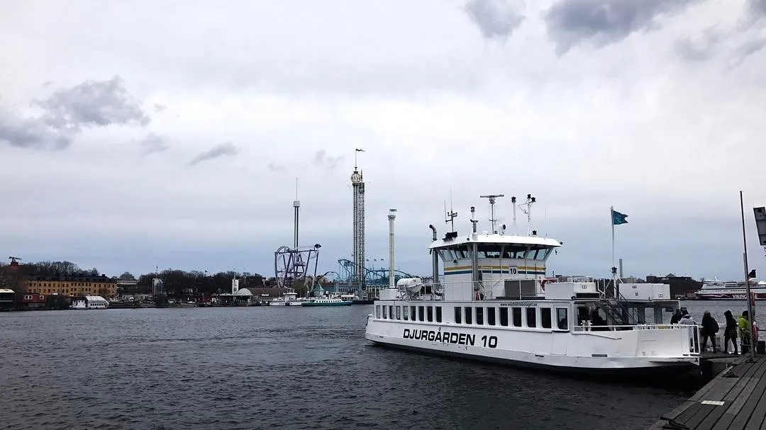 A large white boat labeled DJURGÅRDEN 10 docks on the right, set against a dark harbor and a distant industrial skyline under a heavy, overcast gray sky.