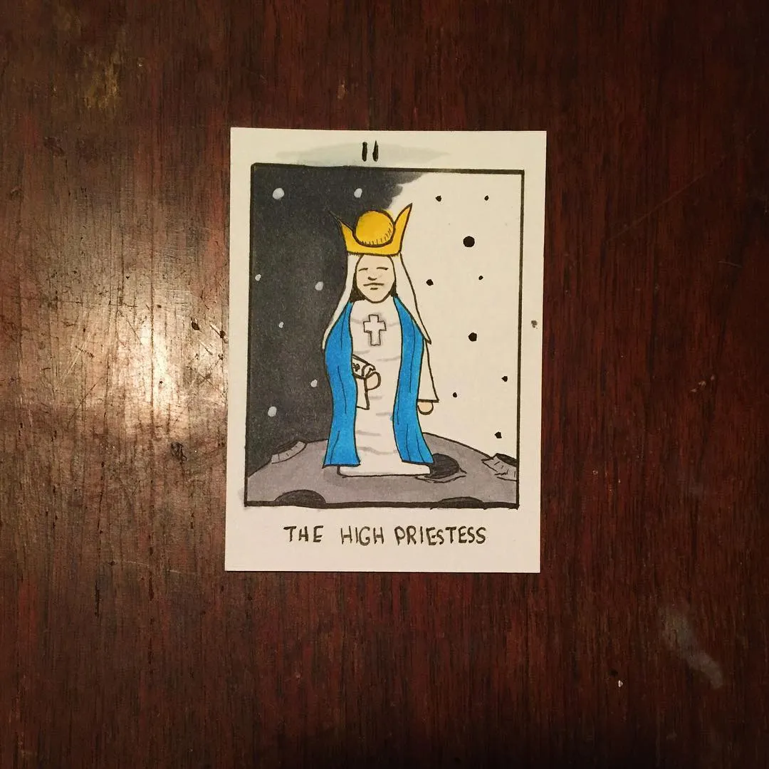 A stylized drawing on white paper depicts a figure in blue and white robes and a gold crown, standing on dark rocks beneath a starry night sky and crescent moon, with the title THE HIGH PRIESTESS printed below.