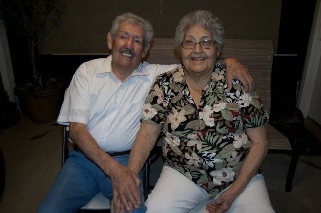 A man in a light blue striped shirt and jeans sits next to a woman in a floral top and white pants, both smiling and posed indoors.