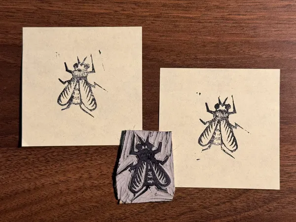 Three stylized drawings of insects are displayed on a dark wood surface: two on light beige paper squares and one on a small, textured piece of wood.
