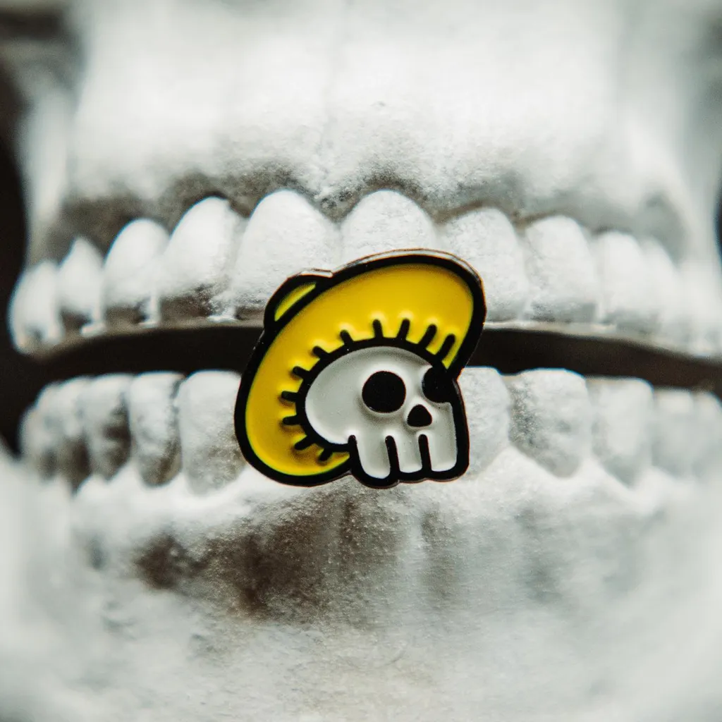 A yellow and black enamel pin featuring a stylized skull with a headband, resting on a close-up surface of white teeth and gums.
