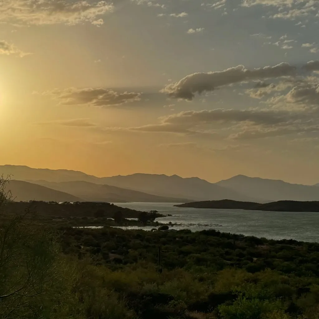 Layered horizontal landscape showing dark foreground vegetation, a calm inlet, and distant mountain ranges under a warm, hazy sky with soft clouds.