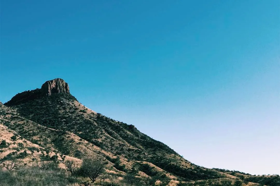 The composition features a steep, scrub-covered mountain rising from the bottom edge against a clear blue sky, culminating in a dark, rocky peak on the left.