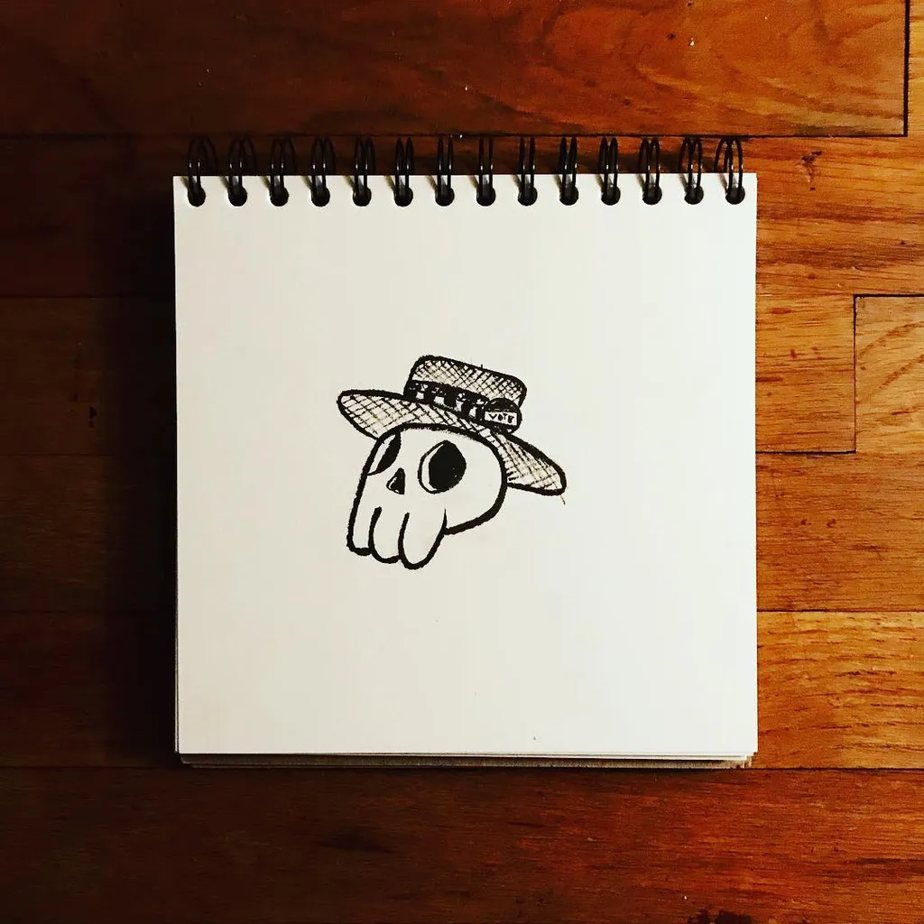 A black ink drawing of a stylized skull wearing a cowboy hat, centered on a spiral-bound sketchbook resting on a wooden surface.