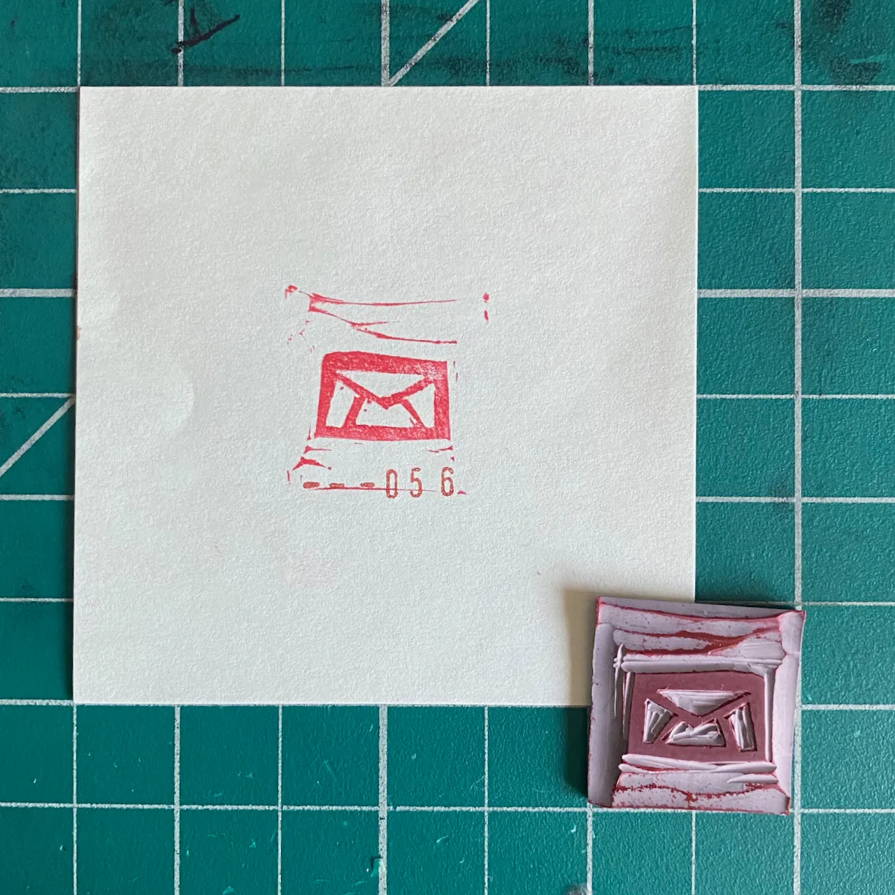 A white square paper on a teal grid displays a red envelope drawing and the numbers 05 6, with a smaller, layered red and white piece featuring a similar design below.
