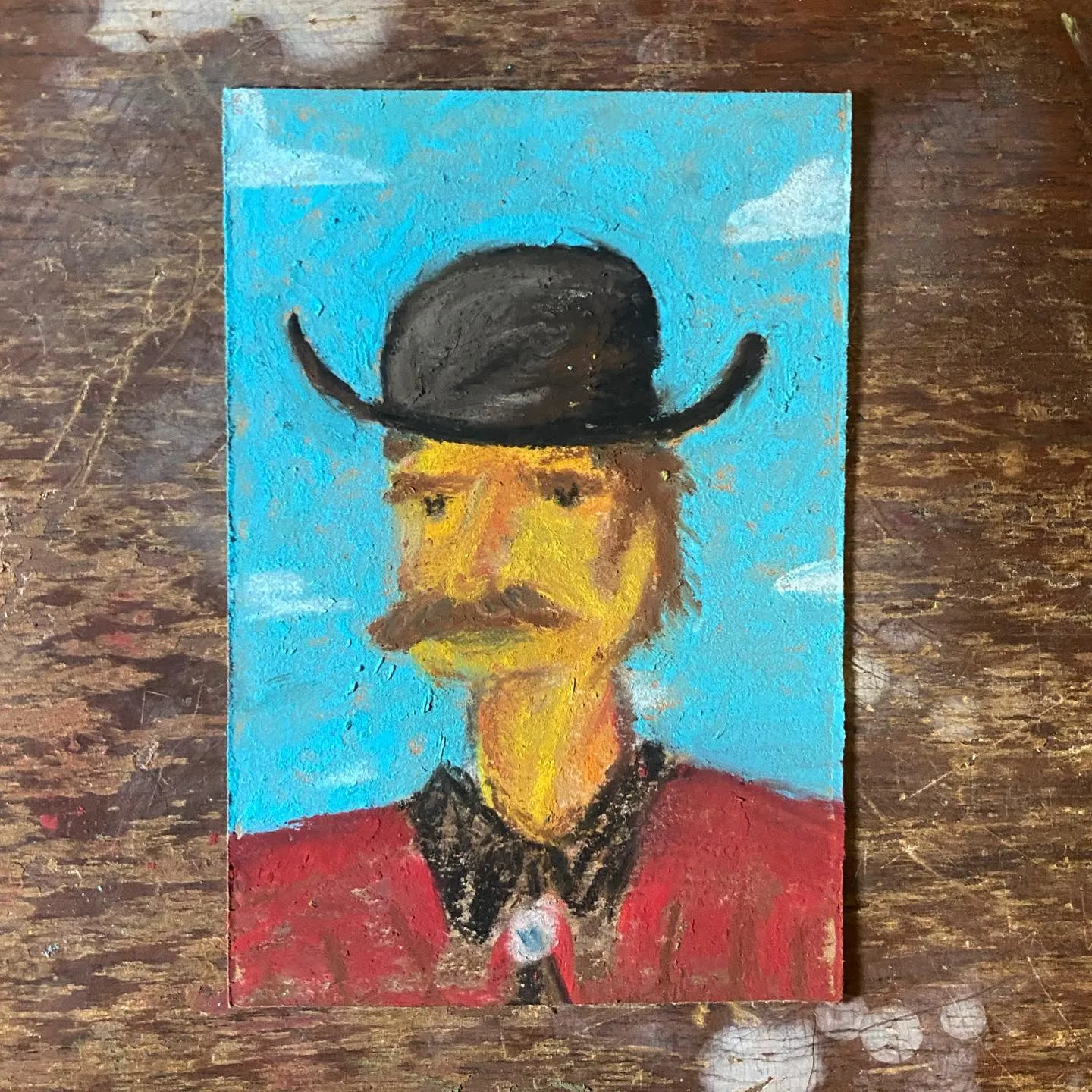 A portrait of a man with a yellow face and mustache, wearing a black hat and red clothing, set against a bright blue background, which rests on a textured brown surface.