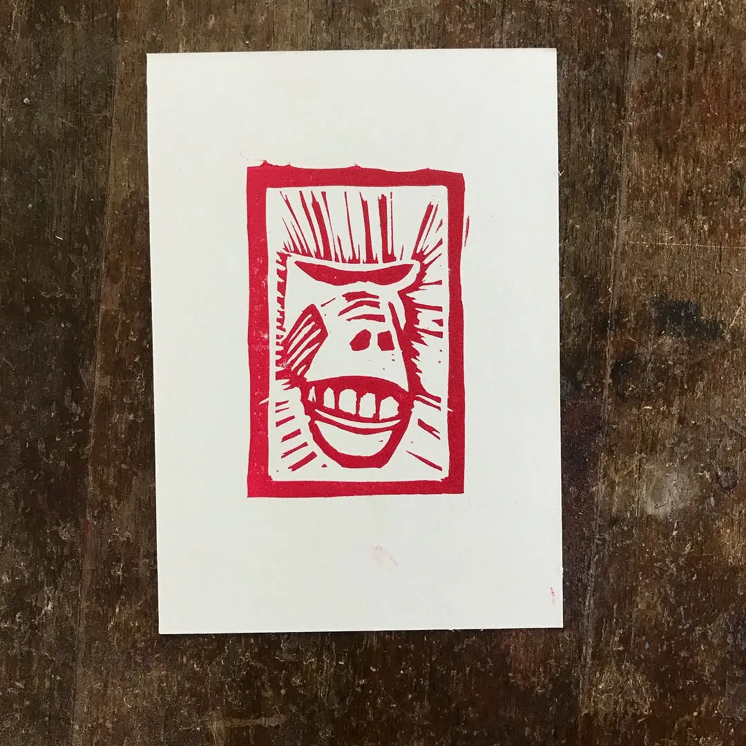 A stark red print on white paper depicts a highly stylized, aggressive face enclosed in a rectangular border, featuring jagged lines and a wide, toothy smile.