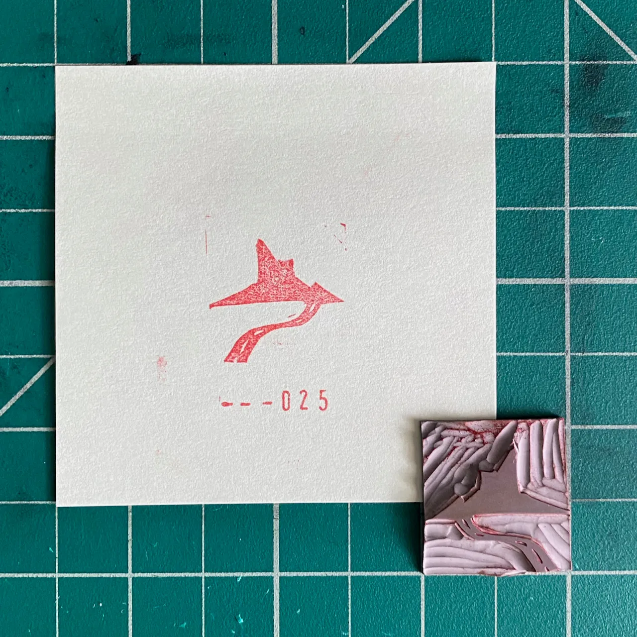 A white card on a teal grid displays a red stylized figure and the number --025, next to a small rectangular piece with marbled pink, white, and gray textures.