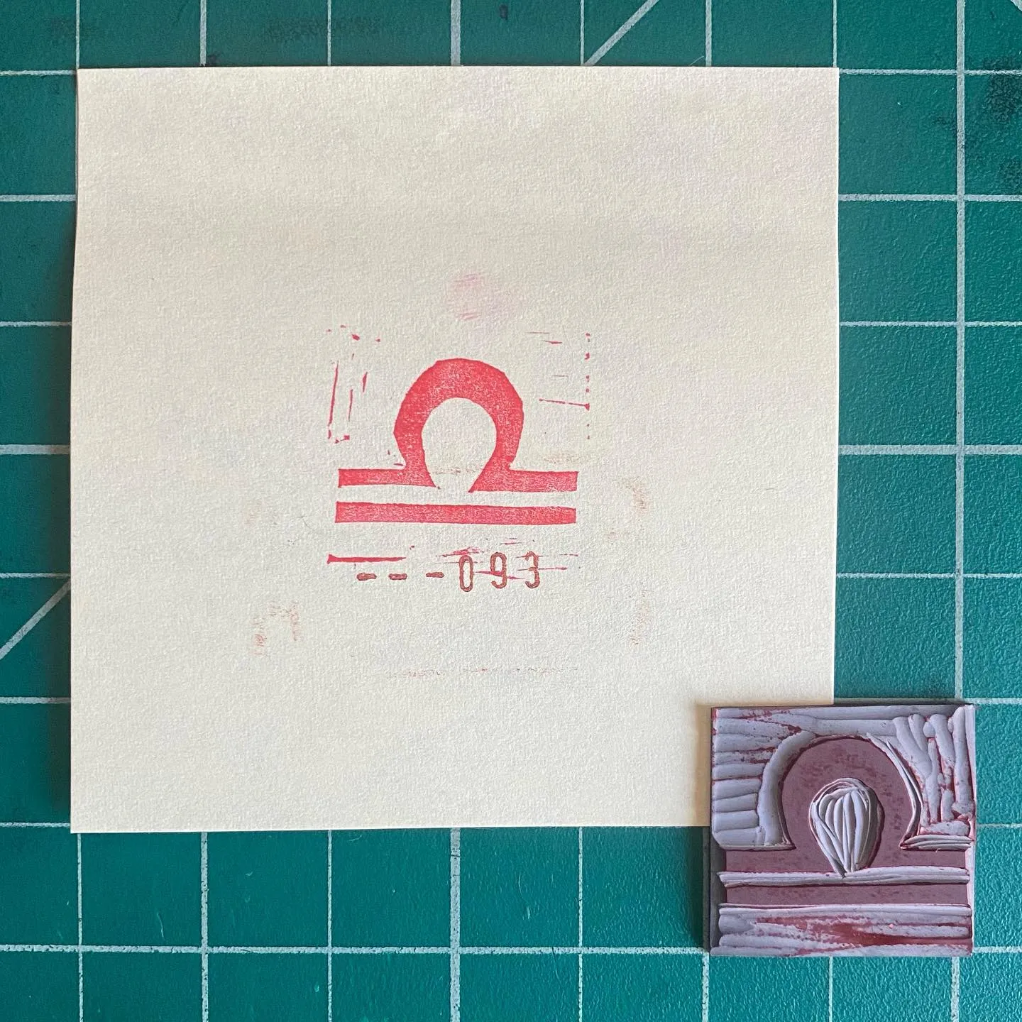 On a teal grid, a cream paper holds a red stylized symbol and the numbers -093, next to a small, layered piece of art with horizontal bands of pink and white and a curved textured design.