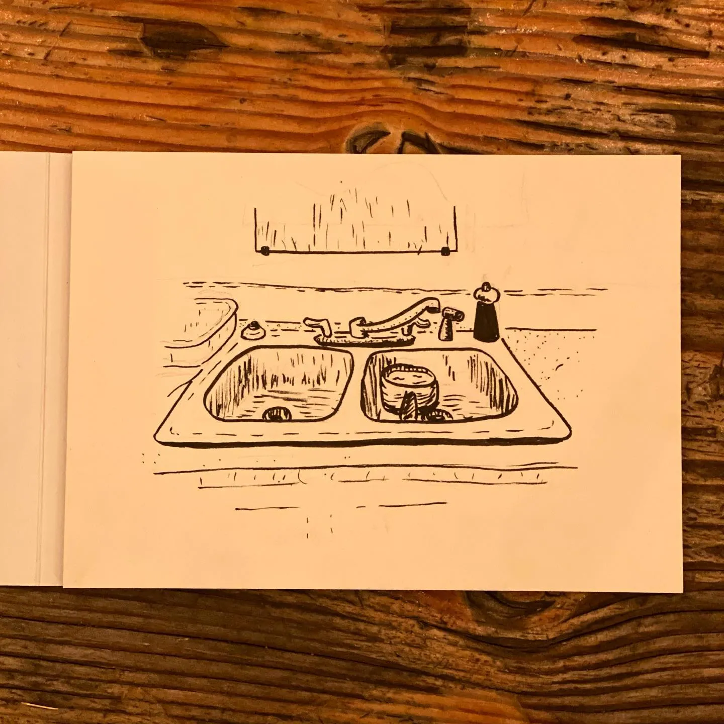 A simple black and white line drawing depicts a rectangular basin divided into two parts, showing sketched objects such as a faucet, soap dispenser, and various curved lines.
